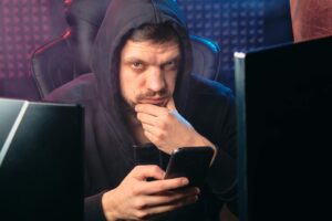 Un homme portant un sweat à capuche noir réfléchit tout en utilisant un smartphone, entouré d'écrans numériques.